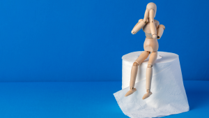 Cilvēka koka figūriņa, kas aizsegusi ar plaukstām sev seju un sēž uz tualetes papīra ruļļa