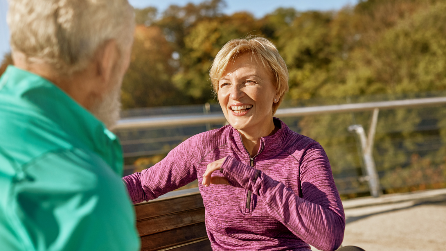 Priecīga sieviete violetā sporta džemperī sēž uz soliņa un sarunājas ar vīrieti. Ārā ir saulains laiks